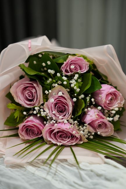 A bouquet of fresh, pastel pink roses with delicate, velvety petals arranged in a rounded, compact style, accented by small white baby's breath flowers and lush green leaves. The bouquet is wrapped in
