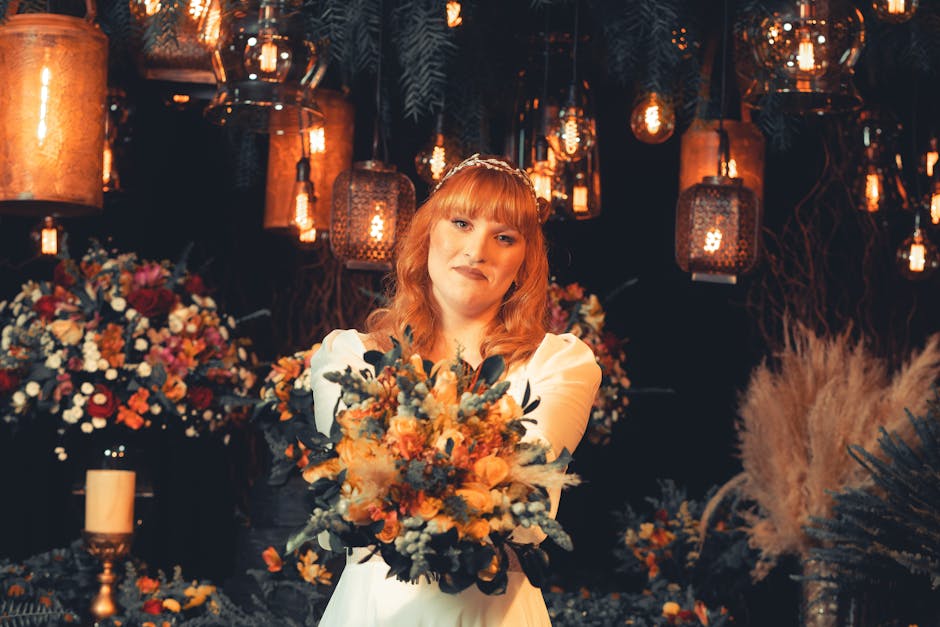 A young woman with ginger hair, dressed in a white dress, is holding a vibrant, mixed bouquet with shades of orange, yellow, blue, and green flowers, including roses, eucalyptus, and dried foliage. Sh