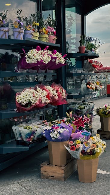 An array of fresh floral arrangements displayed on metal shelves inside a floral shop, with bouquets wrapped in colorful paper and ribbons, featuring white, pink, purple, and yellow flowers including 