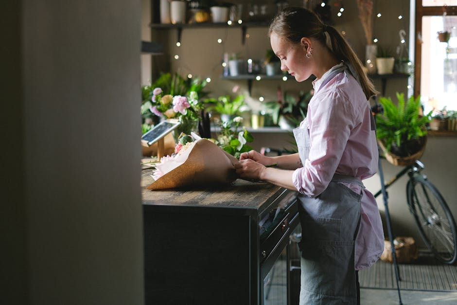 A young female florist in a cozy, well-lit flower shop arranging a bouquet of fresh flowers on a wooden counter. She is wearing a light pink shirt and a gray apron, with her hair tied back. The bouque