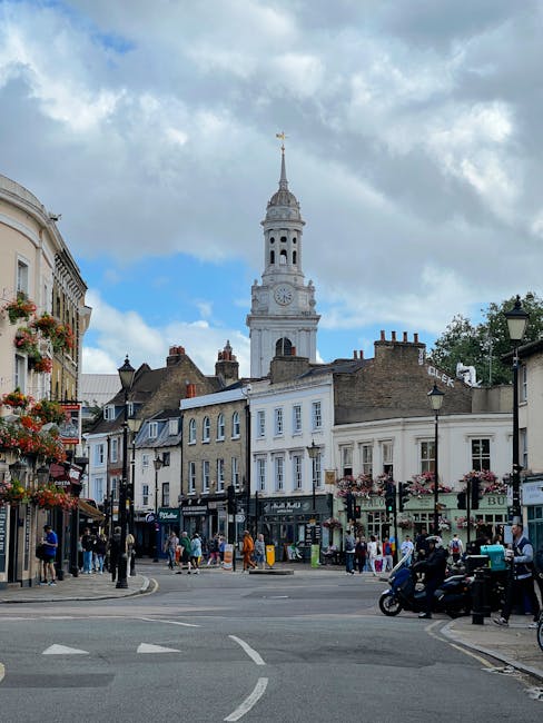 A bustling street scene in a historic UK town with a prominent white clock tower topped with a weather vane in the background. The foreground features a variety of shops and businesses housed in tradi