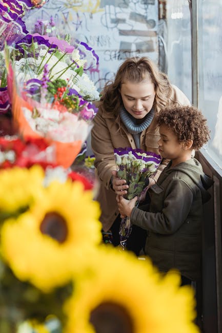A young woman with light brown, wavy hair dressed in a beige coat and a blue turtleneck is smiling warmly as she hands a small bouquet of fresh purple and white flowers to a curly-haired boy wearing a